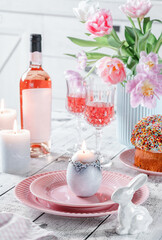 Easter holidays table, pink wine in glasses, Easter cake, color eggs, served plates, flowers tulips, candles on white background for festive dinner at home. Holiday concept, close up