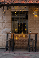 Two empty small tables and chairs in front of the window with light decorations of a small cozy cafe. Shot at pedestrian El Conde street in Santo Domingo historical center, Dominican Republic. - obrazy, fototapety, plakaty