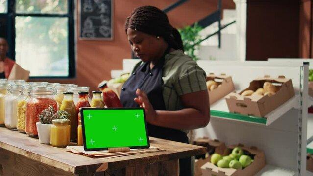 Tablet display showing greenscreen in local organic shop, screen running isolated chromakey layout next to women chatting about chemicals free goods. Blank copyspace template. Tripod shot.