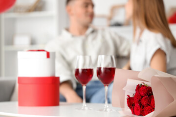 Beautiful young couple with glasses of wine, bouquet and gift box celebrating Valentine's Day at home
