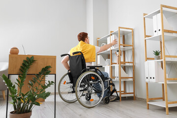 Young man in wheelchair taking folder from shelf at office, back view