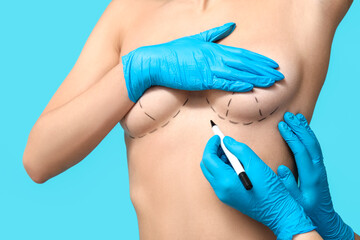 Doctor drawing marks on female breast before cosmetic surgery operation against blue background