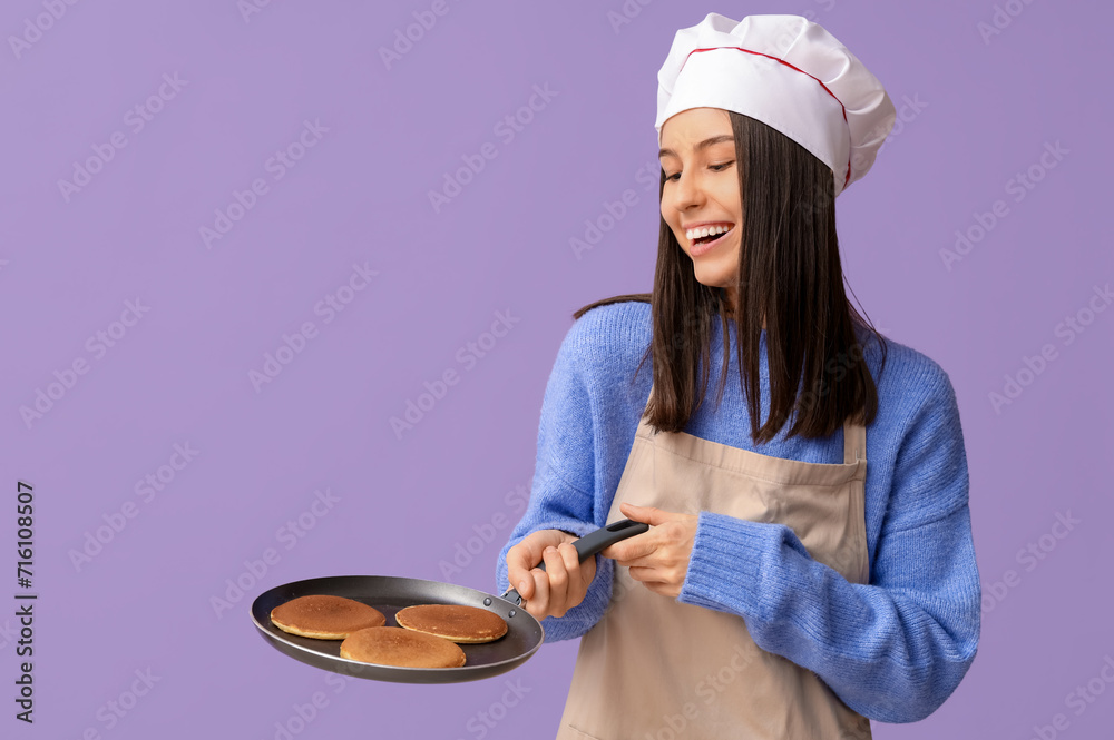 Wall mural Young female chef holding frying pan with tasty pancakes on lilac background