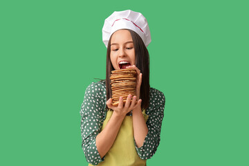 Young female chef eating tasty pancakes on green background