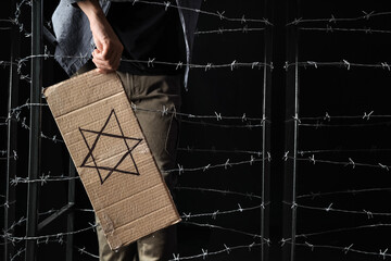 Young Jewish man holding paper with David star behind barbed wire on black background....