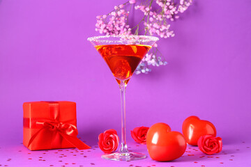 Glass of tasty cocktail with hearts, roses and gift on purple background. Valentine's Day celebration