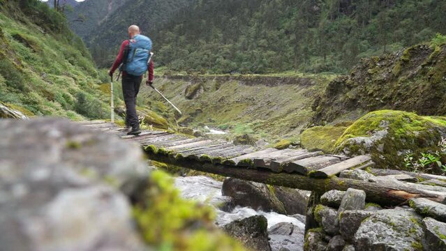 Man with backpack and trekking poles crossing mountain creek wooden bridge during Makalu Barun National Park trek in Nepal. Mountain hiking and active people concept image.