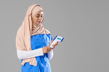 Female Muslim medical intern with infrared thermometer on light background