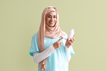 Female Muslim medical intern pointing at pill bottle on green background