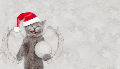 Happy cat wearing red santa hat holds big snowball while lying on snow. Empty space for text