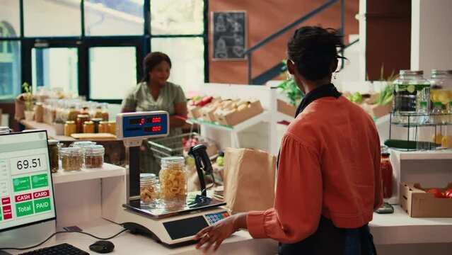 Local merchant placing groceries on scale at cash register, ensuring good pricing value for customer at farmers market. African american storekeeper serving client, eco friendly. Handheld shot.