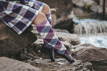 Woman in purple and blue tartan skirt and long woolen socks sitting on rocks beside waterfall in...