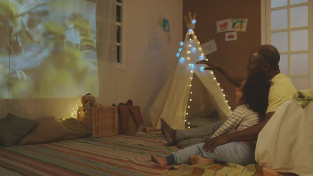 Side full footage of African American father talking about bees sitting with sleepy son on floor in decorated room with teepee while watching educational video with projector in evening