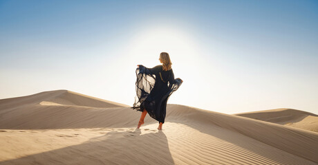 Fototapeta na wymiar Desert adventure. Young arabian Woman posing in traditional Emirati dress abaya in sanddunes of UAE desert at sunset. The Dubai Desert Conservation Reserve, United Arab Emirates.