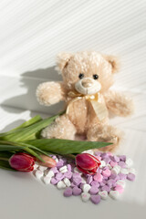Background for Valentine's Day. Assorted pink and white heart shaped candies with printed messages of love, pink tulips and beige cute Teddy bear on a white background. Gift for Women's Day, March 8
