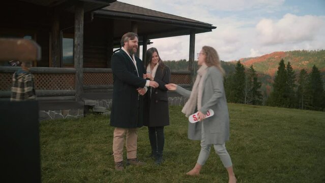 Professional real estate agent gives keys and congratulates couple with their purchase. Realtor puts sold sticker on for sale sign. Young family buys modern house in mountains with amazing view.