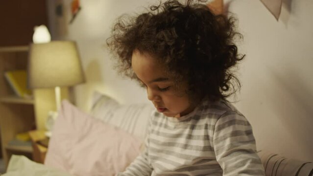 Side tilt shot of five-year-old African American boy in pajamas sitting in bed before sleep and looking at dinosaur cartoons paging through comic book before going to sleep