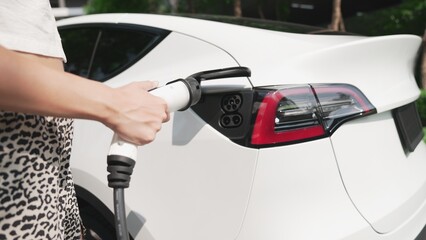 Young woman travel with EV electric car charging in green sustainable city outdoor garden in...