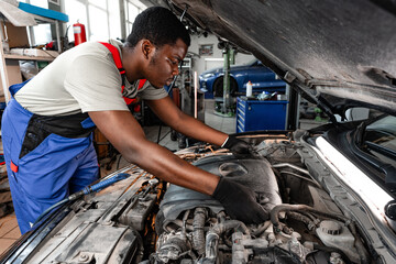 Young African male mechanic repairs car in garage close up - obrazy, fototapety, plakaty