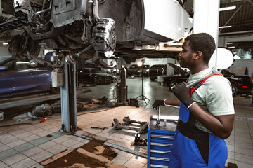 African male auto-mechanic repairing car brakes under the car in auto service - obrazy, fototapety, plakaty