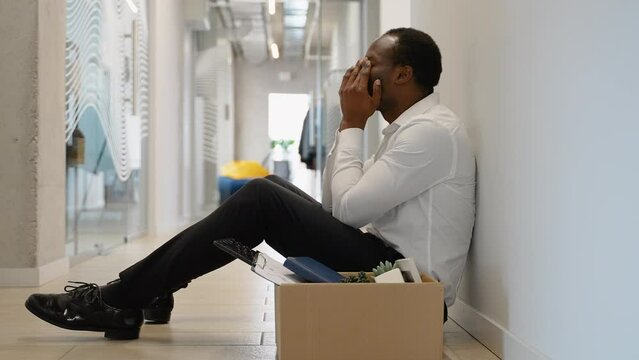Fired african american worker sitting frustrated and upset in the hallway near office with dismissal box