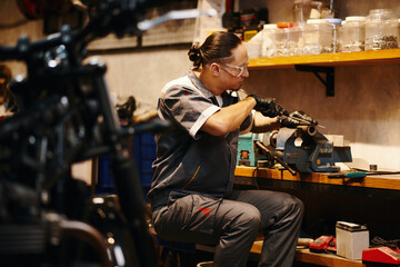 Repairman in protective googles filing motorcycle detail at his workbench - obrazy, fototapety, plakaty