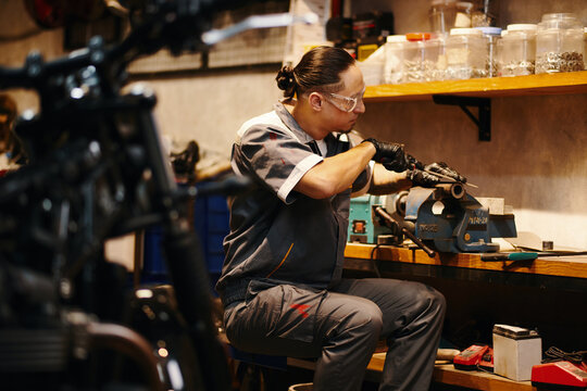 Repairman in protective googles filing motorcycle detail at his workbench