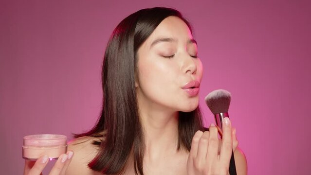 Young Asian girl applying make up powder on face against pink background