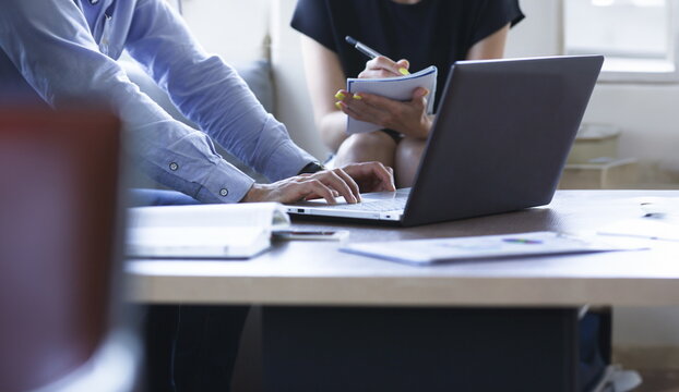 Two business people are comparing information one is working behind a laptop business woman is making notes on a pen in her notebook in a cafe at a table.