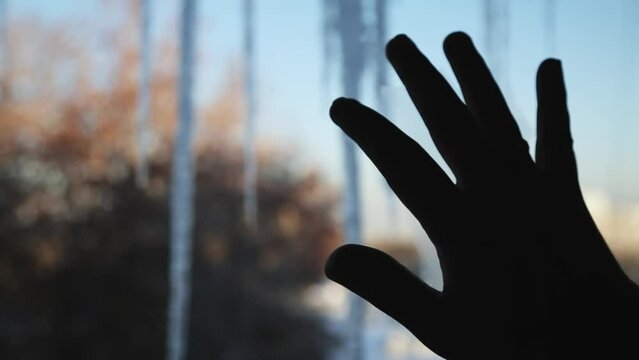 A young man's hand touches icicles hanging on a window against the background of a winter city.