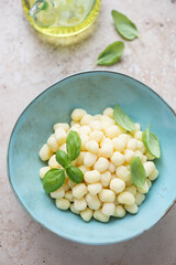 Turquoise bowl with raw italian chicche di patate and fresh green basil, vertical shot on a beige granite surface, elevated view