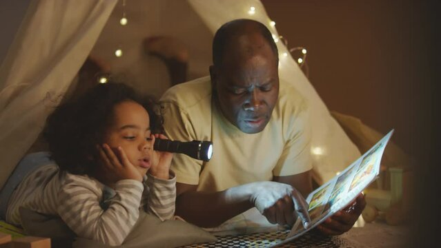 Panning shot of African American kid in pajamas with flashlight lying in play handmade wigwam decorated with garland with father who reading comics in bedroom at night