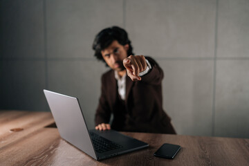 Portrait of serious successful businessman in suit looking at camera and pointing finger forward calling to action. Front view of business man working on laptop sitting at table modern office.