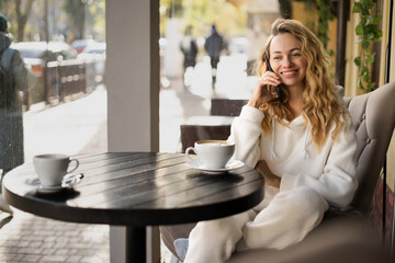 Happy blonde woman speaking with her friend by phone while relaxing in cozy coffee shop, talking on...