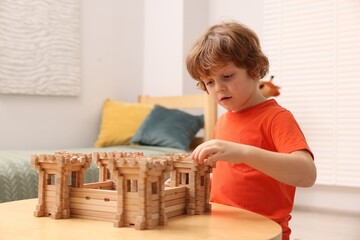 Cute little boy playing with wooden fortress at table in room. Child's toy