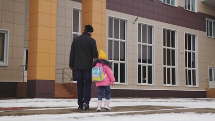 little girl holds father hand, happy family, little child kid backpack walks school snow, dad escorts kid child daughter school, cold winter, suitcase man walks happily with daughter white snow road,