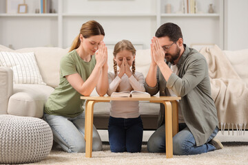 Girl and her godparents praying over Bible together at table indoors