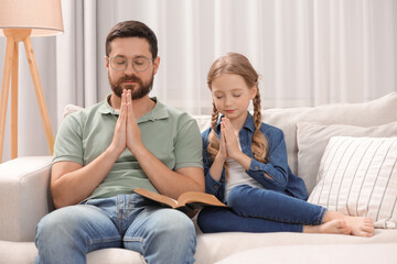 Naklejka na ściany i meble Girl and her godparent praying over Bible together on sofa at home