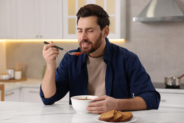 Man eating delicious tomato soup at light marble table in kitchen