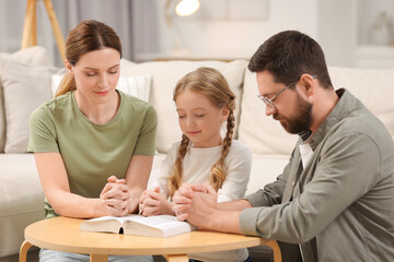 Girl and her godparents praying over Bible together at table indoors