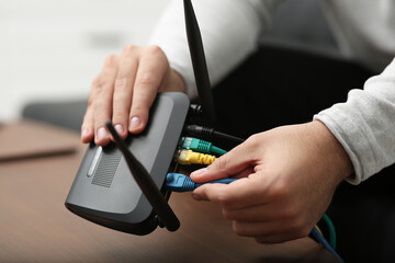 Man inserting cable into Wi-Fi router at wooden table indoors, closeup