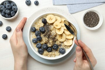 Woman eating tasty oatmeal with banana, blueberries and chia seeds at white wooden table, top view
