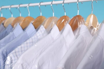 Dry-cleaning service. Many different clothes in plastic bags hanging on rack against light blue background, closeup