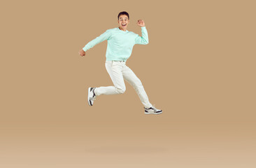 Fototapeta na wymiar Happy cheerful excited young man or teenage boy in casual mint sweatshirt and white pants running and jumping high in air isolated on light brown color background