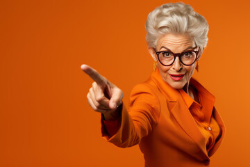 Older Woman with her finger pointing on a isolated orange background. Senior woman pointing with hand and finger to the side looking at the camera. - obrazy, fototapety, plakaty