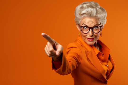 Older Woman with her finger pointing on a isolated orange background. Senior woman pointing with hand and finger to the side looking at the camera.