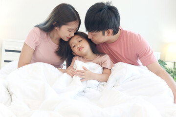 Happy Asian family has fun in bedroom. Father, mother daughter cover body with blanket during lying together on white bed. Parents kiss kid child girl has good memory at home. parenthood and childhood