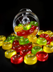 A lot of candy canes are scattered from an overturned glass goblet. Multi-colored candies in a glass on a black background.