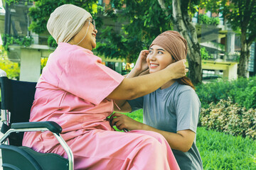 Elderly happy mother, wearing headscarf is suffering from cancer and is sitting wheelchair with daughter sitting next to her showing her love she wears cloth hat encourage her and wish her well.