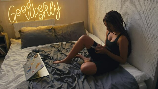 High angle footage of young African American woman sitting with phone in bed and vaping near laptop in modern grunge loft apartment with neon sign Good Vibes Only on wall